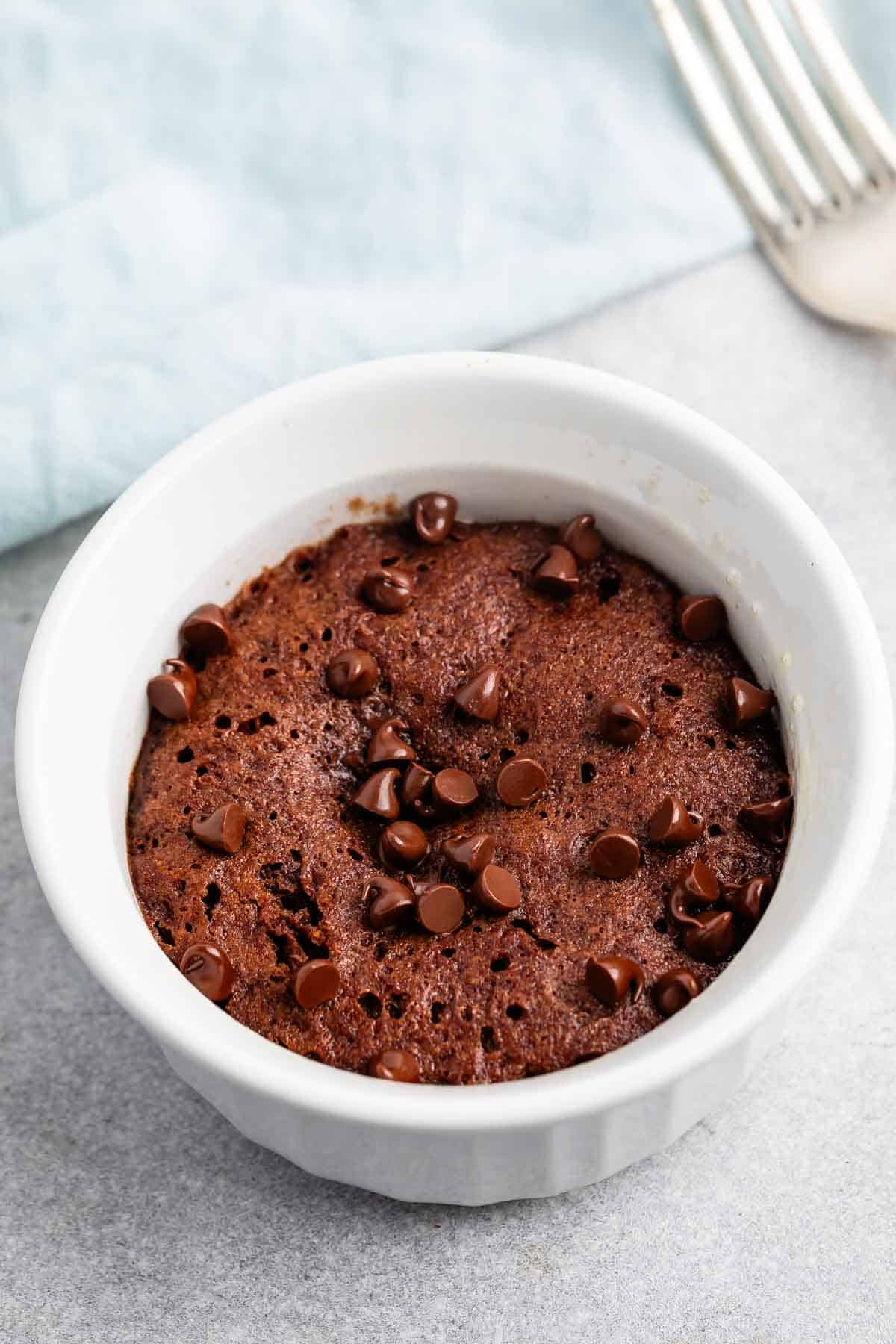 A chocolate mug cake topped with chocolate chips in a white ramekin, placed on a gray surface. A silver fork is partially visible in the background, and a light blue cloth is nearby.