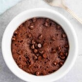 A chocolate mug cake topped with chocolate chips in a white mug on a gray stone surface. A fork is placed nearby.