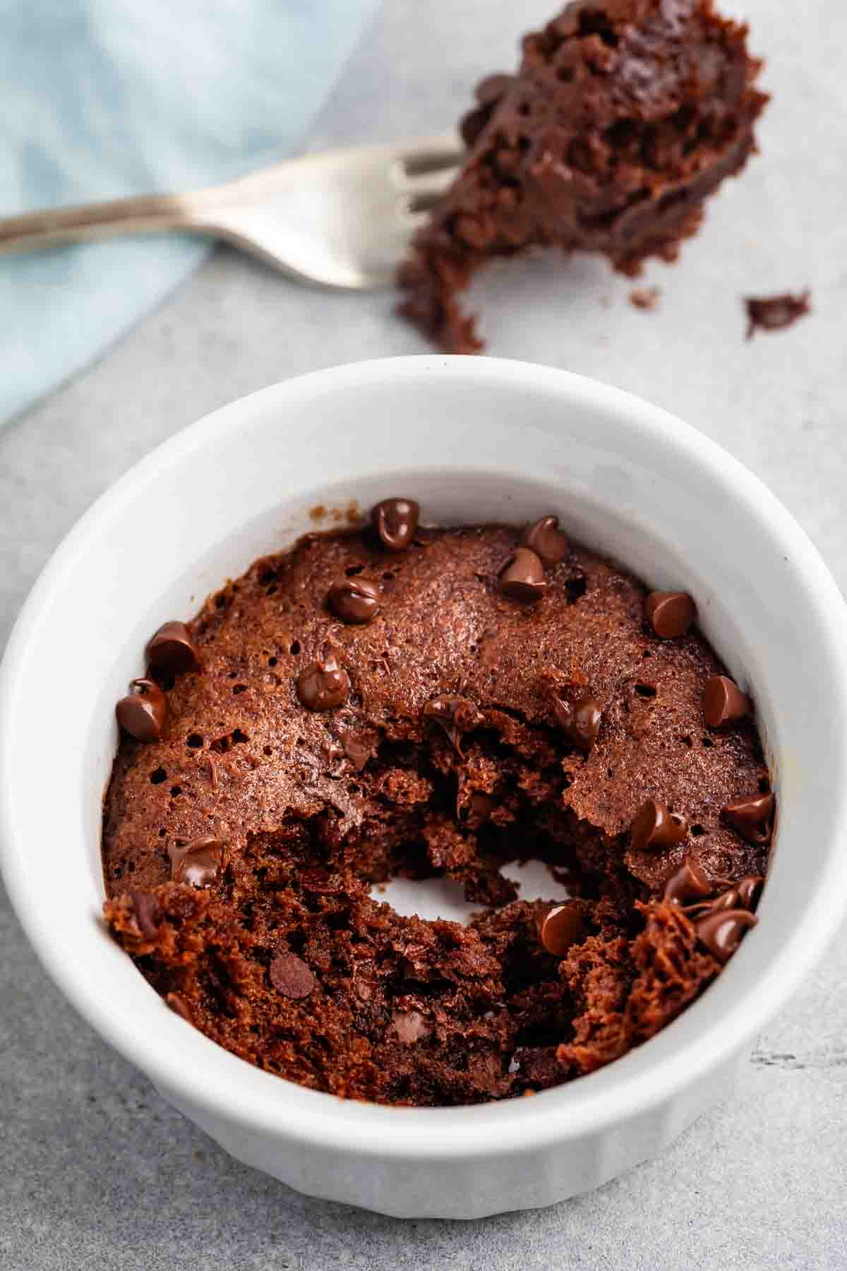 A partially eaten chocolate mug cake with scattered chocolate chips, served in a white ramekin. A fork with cake crumbs rests nearby on a light gray surface.