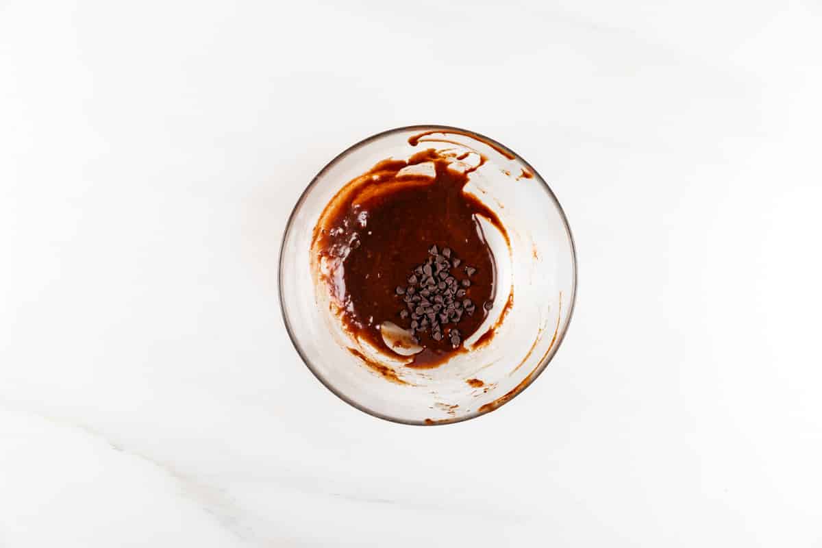 A clear glass bowl containing partially mixed brown batter with chocolate chips on top, set on a white marble surface.