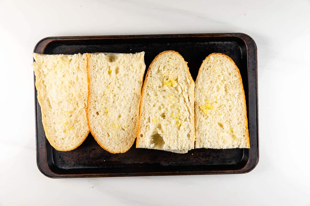 Two sliced baguettes with butter spread on each half are placed on a dark baking tray. The tray is on a white surface.