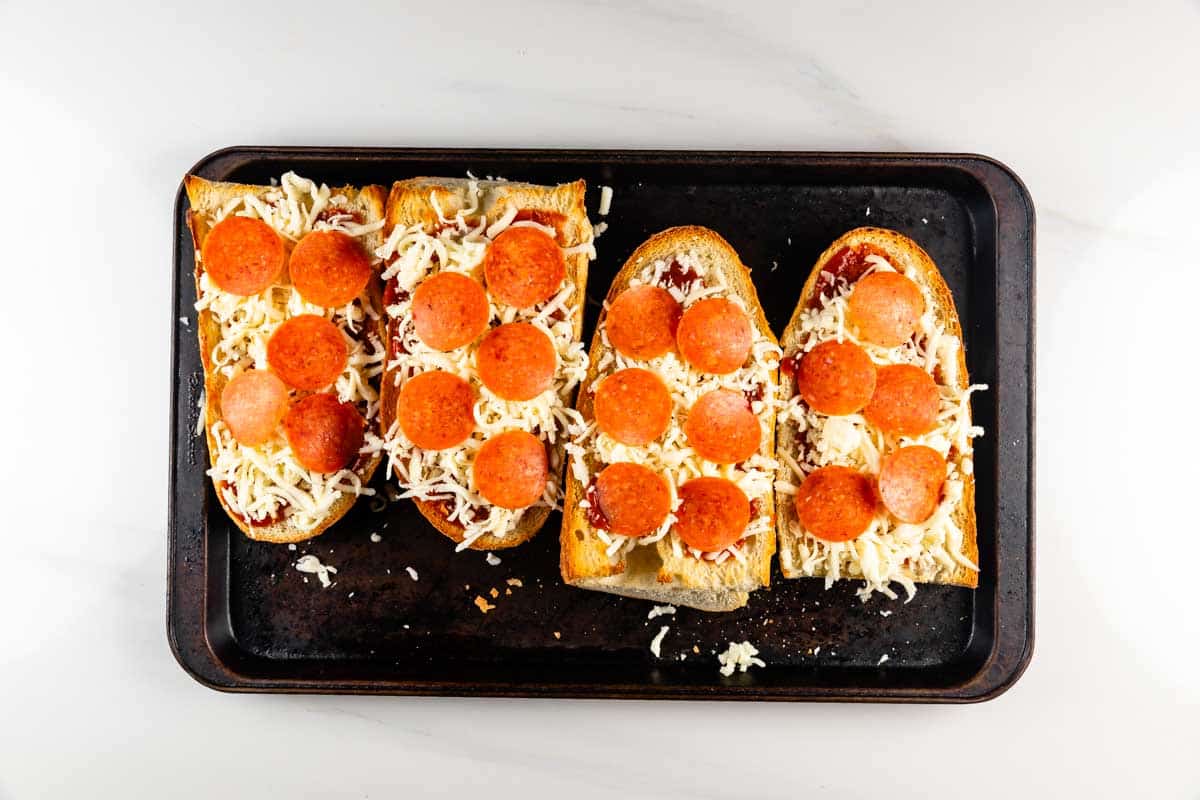 Four slices of French bread on a baking sheet, topped with tomato sauce, shredded cheese, and pepperoni slices, ready to be baked.