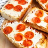 Slices of bread topped with melted cheese and pepperoni are arranged on a wooden surface. A striped cloth is partially visible in the background.