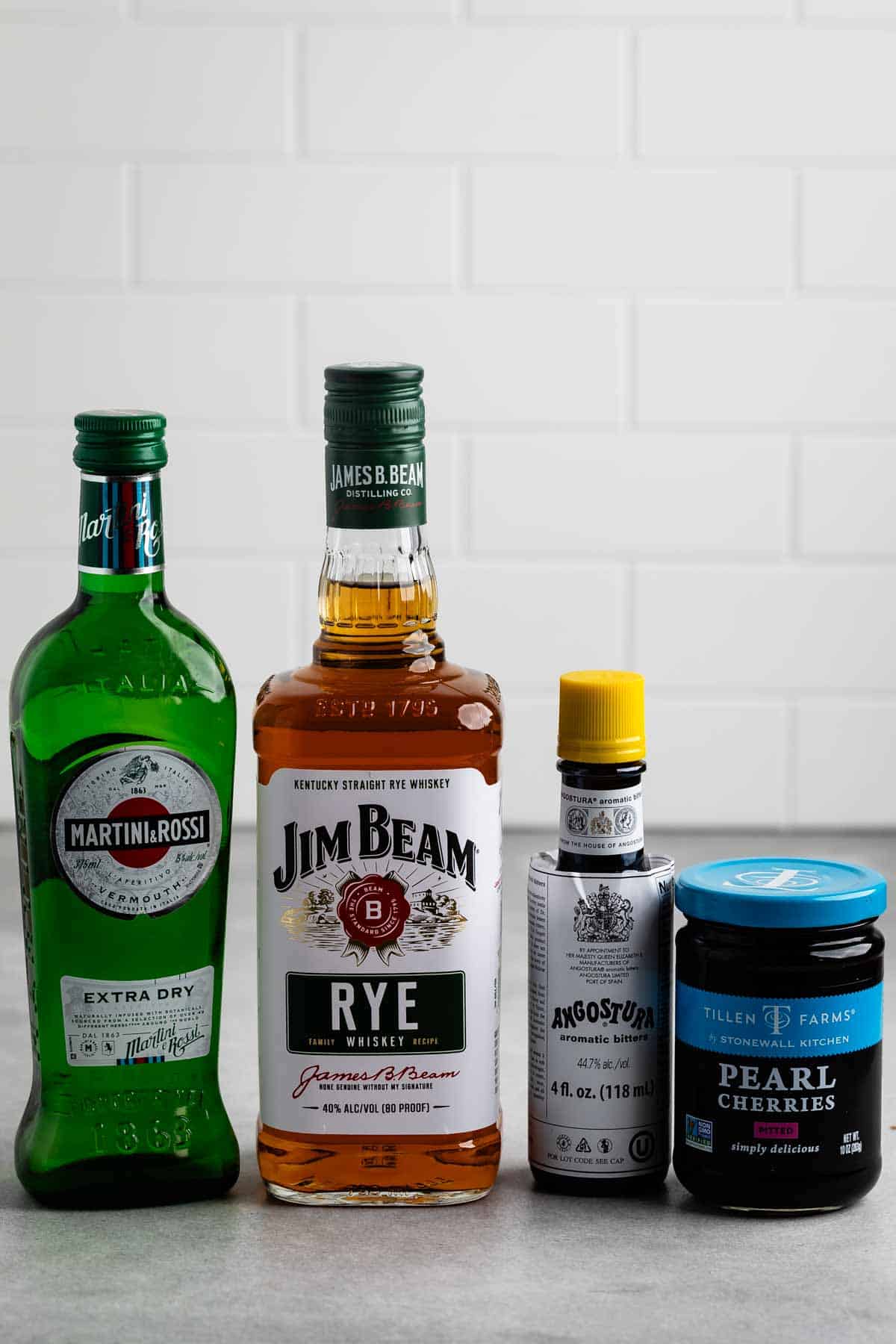 Bottles of Martini & Rossi extra dry vermouth, Jim Beam rye whiskey, Angostura bitters, and a jar of Tillen Farms pearl cherries are lined up on a counter with a white tile background.