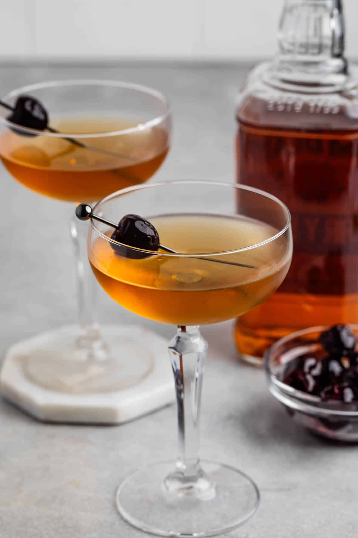 Two cocktails in coupe glasses with cherries on skewers, a bottle of amber liquid in the background, and a small bowl of cherries, set on a light gray surface.