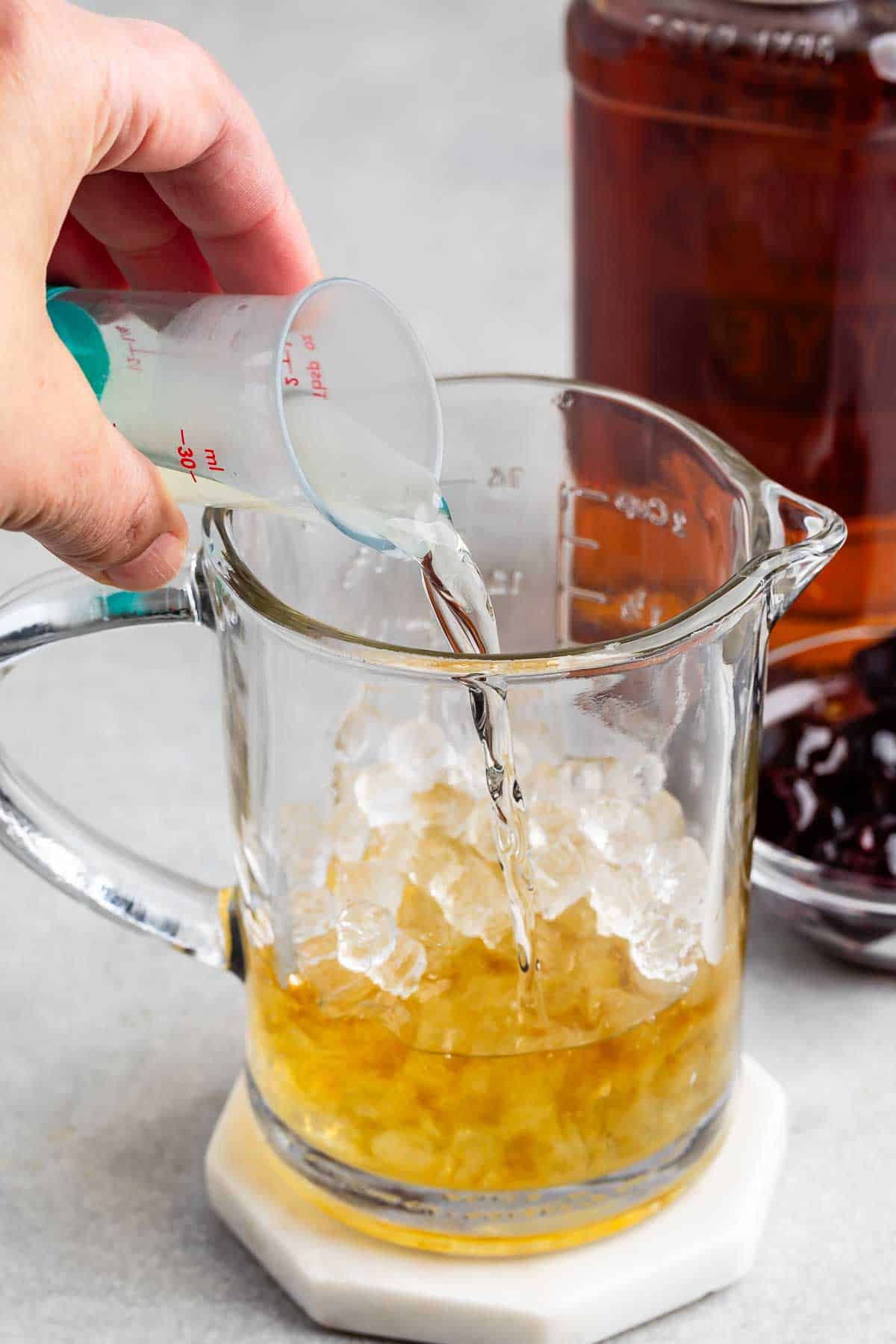 A hand pours liquid from a small measuring cup into a glass pitcher filled with ice and amber liquid. A jar with a brown drink and a bowl of cherries are in the background on a gray surface.