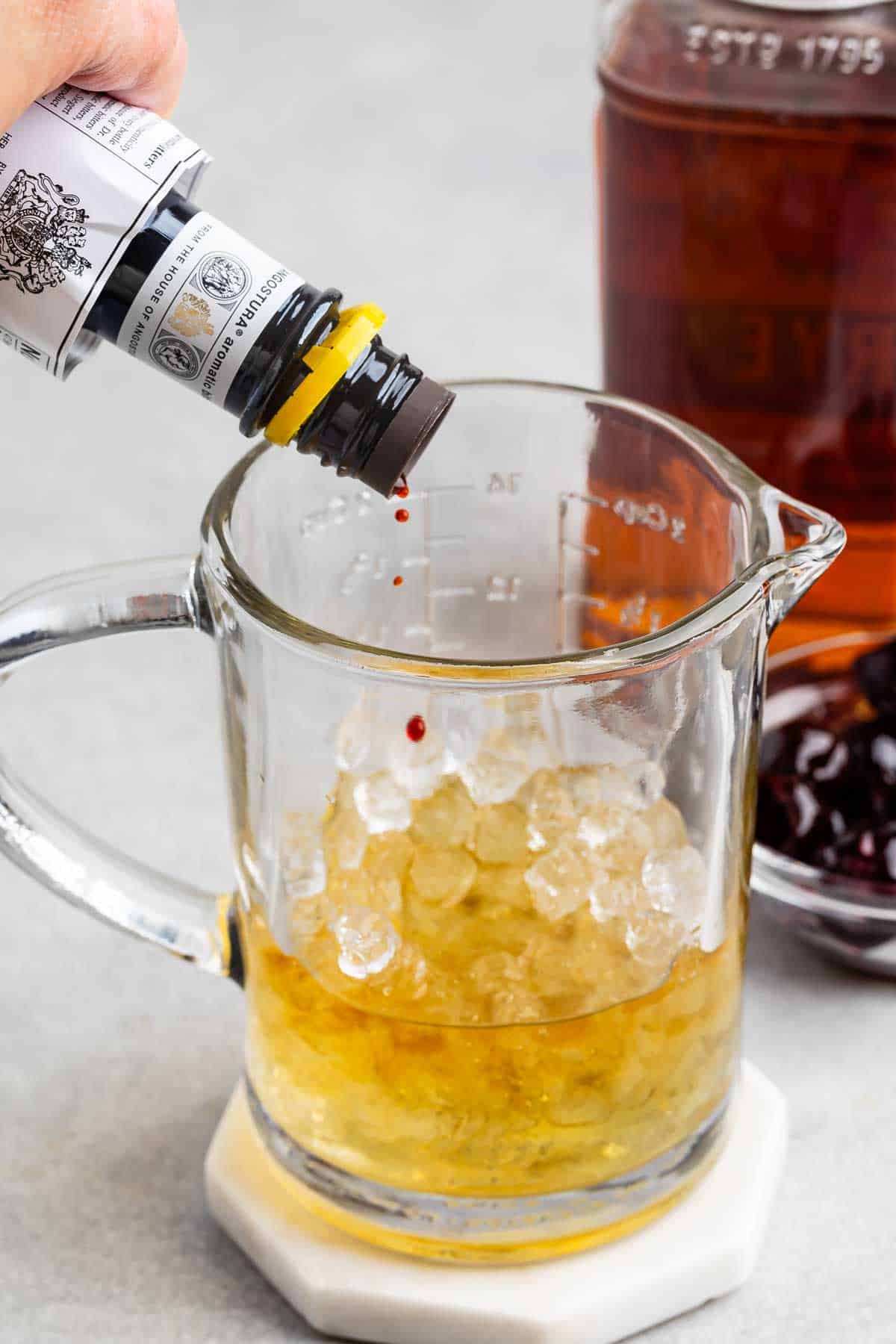 A person adds drops of bitters to a glass measuring cup filled with ice and liquid. In the background, theres a bottle of amber liquid and a bowl of dark cherries on a light countertop.