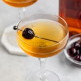 Two elegant cocktail glasses filled with a golden liquid, each garnished with a dark cherry on a skewer. A glass jar of cherries is nearby, set on a light surface.