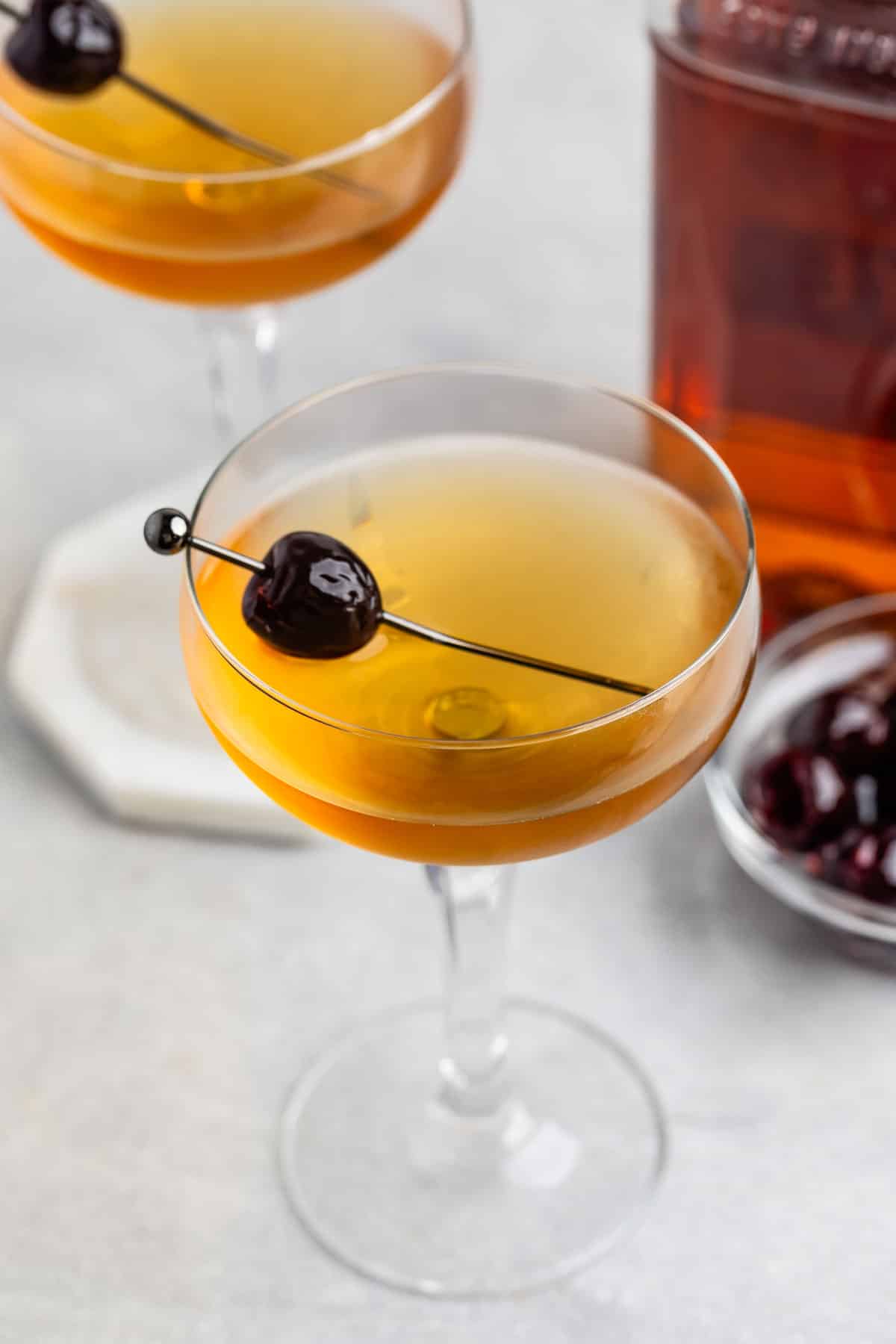 Two elegant cocktail glasses filled with a golden liquid, each garnished with a dark cherry on a skewer. A glass jar of cherries is nearby, set on a light surface.