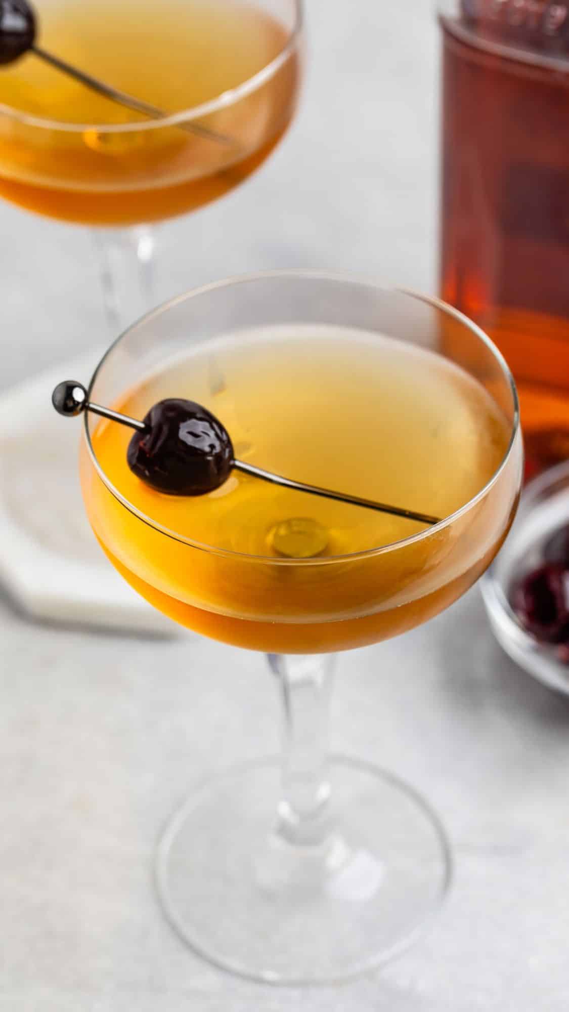 A cocktail in a coupe glass garnished with a dark cherry on a skewer. Another similar glass is in the background, along with a bowl of cherries and a bottle. The setting is on a light surface, creating a sophisticated ambiance.