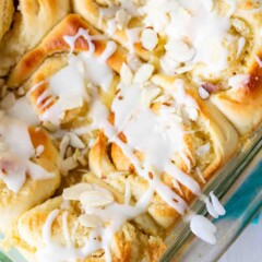 Close-up of freshly baked cinnamon rolls in a glass dish, topped with white icing and sliced almonds. The rolls are golden brown, with a soft and fluffy texture visible under the glaze.