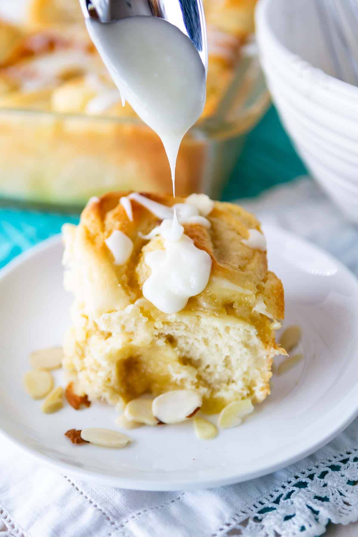 A slice of cinnamon roll on a white plate, topped with sliced almonds. A spoon drizzles white icing over the roll. A baking dish and a white bowl are in the background on a turquoise surface.