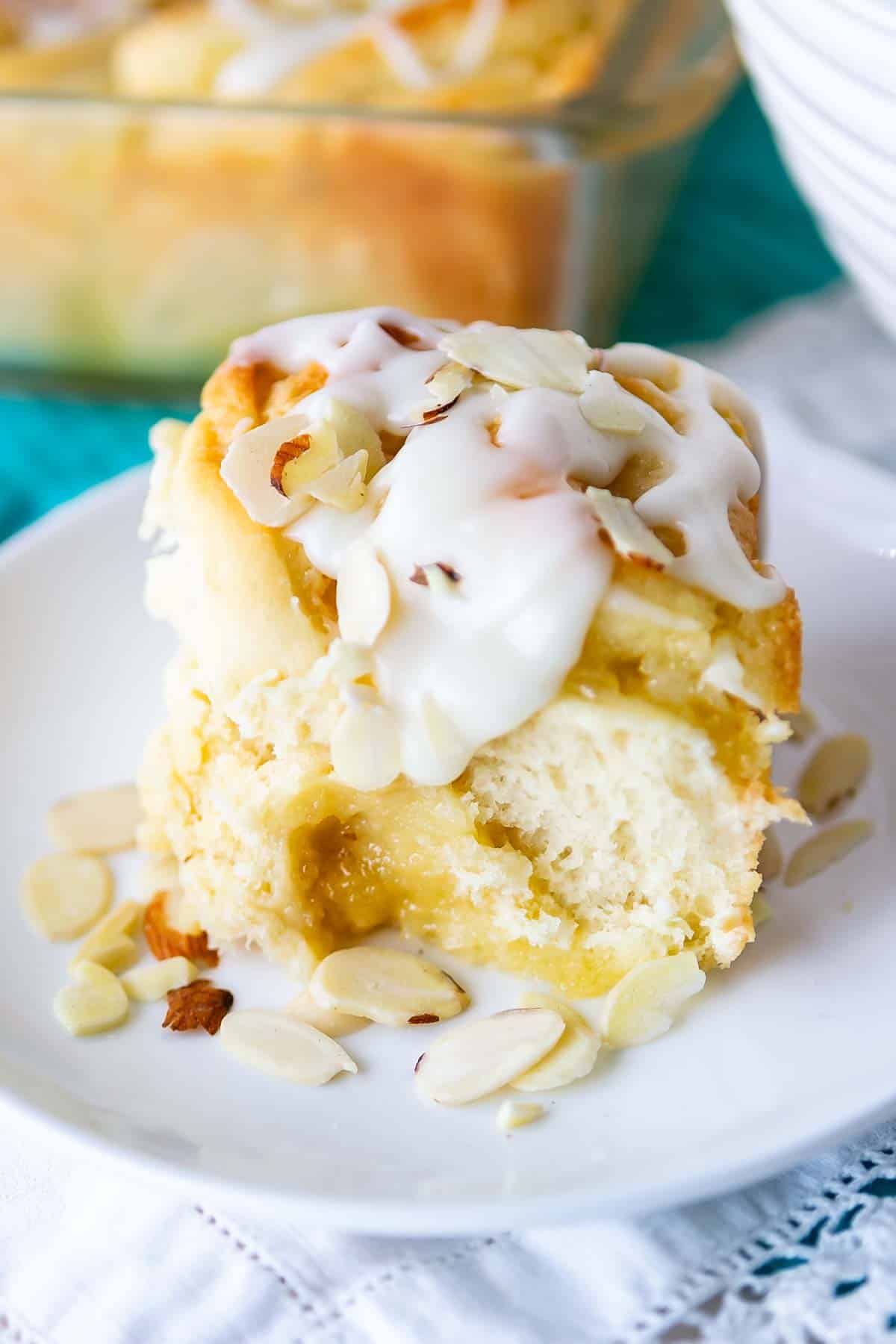 A close-up of a lemon sweet roll topped with creamy icing and sliced almonds on a white plate. A glass baking dish with more rolls is in the background. The setup is on a turquoise and white surface.