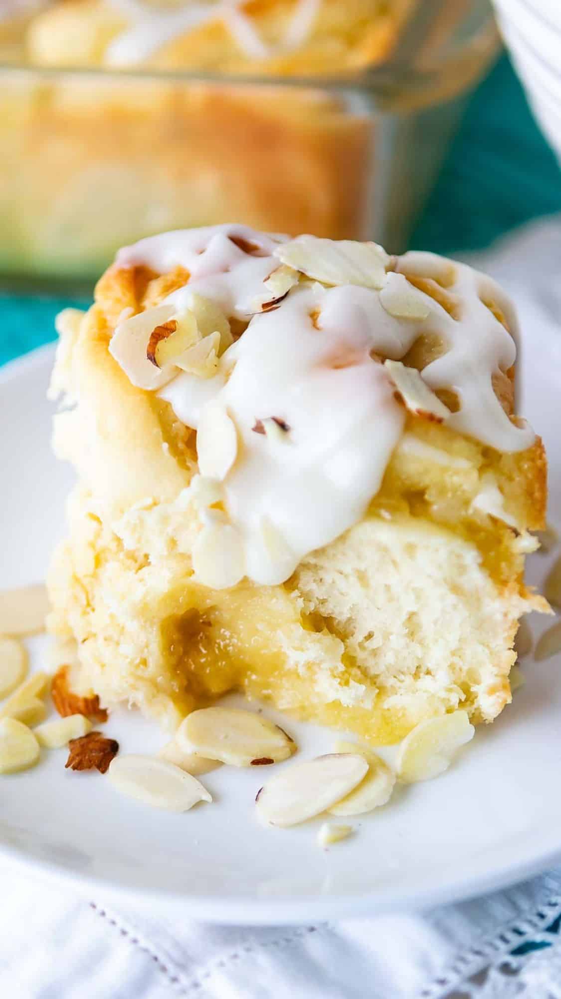 A close-up of a cinnamon roll topped with white icing and almond slices, on a white plate. The roll is fluffy and golden, with a creamy filling oozing out. More rolls can be seen in a glass dish in the background.