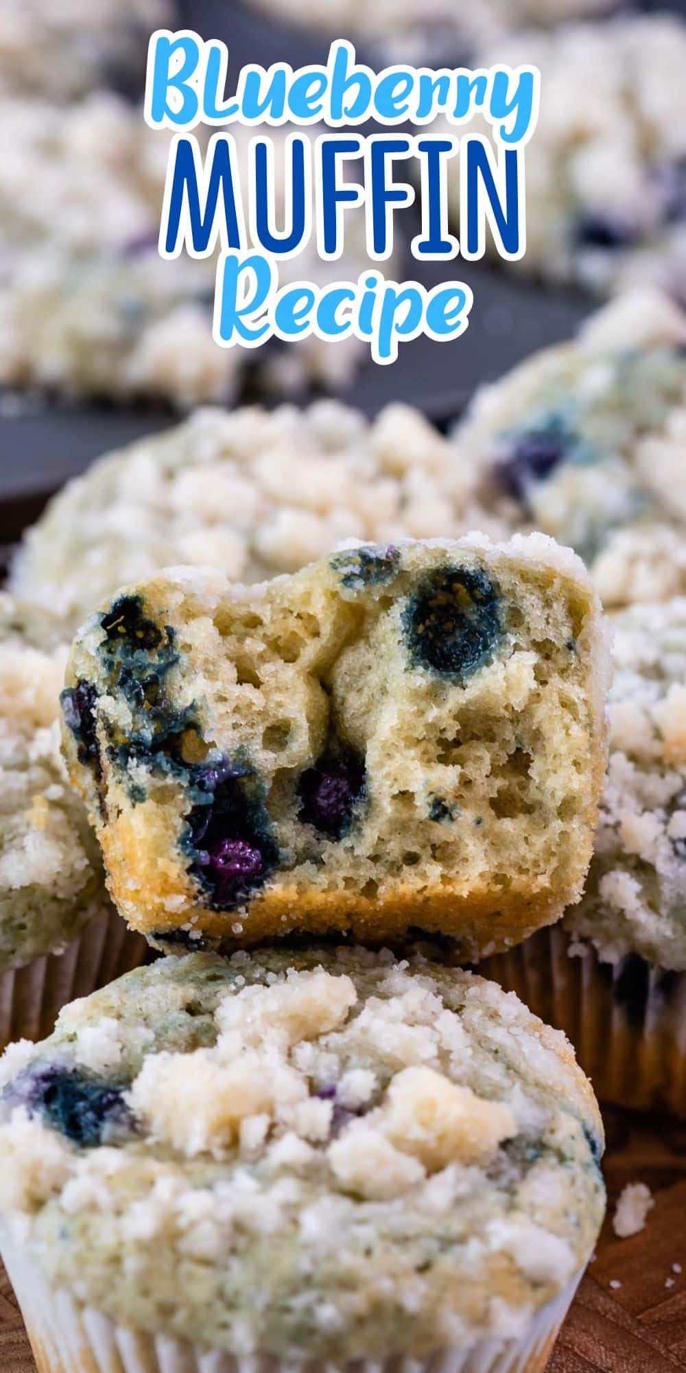 Close-up of blueberry muffins with crumb topping. One muffin has a bite taken out, revealing moist interior with blueberries. Text above reads Blueberry Muffin Recipe.