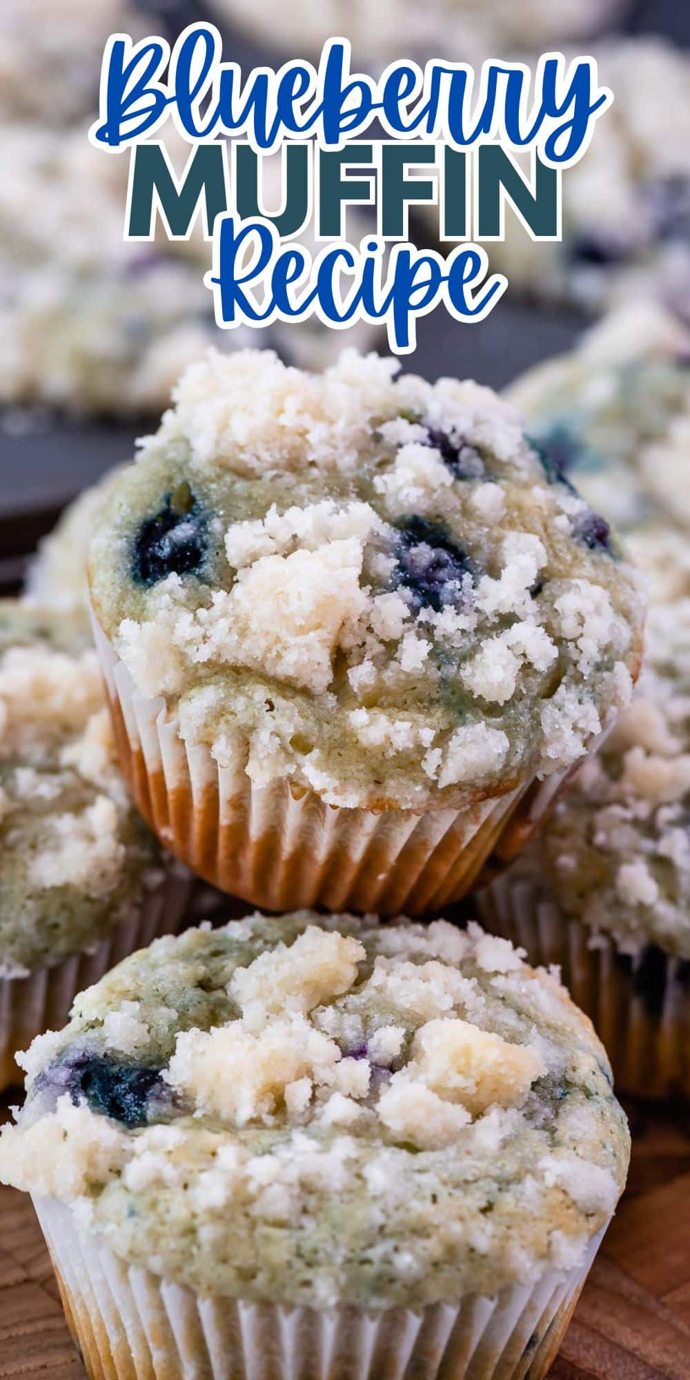 Close-up of freshly baked blueberry muffins topped with crumbly streusel. The top muffin is highlighted with the text Blueberry Muffin Recipe above it. The background shows more muffins on a wooden surface.