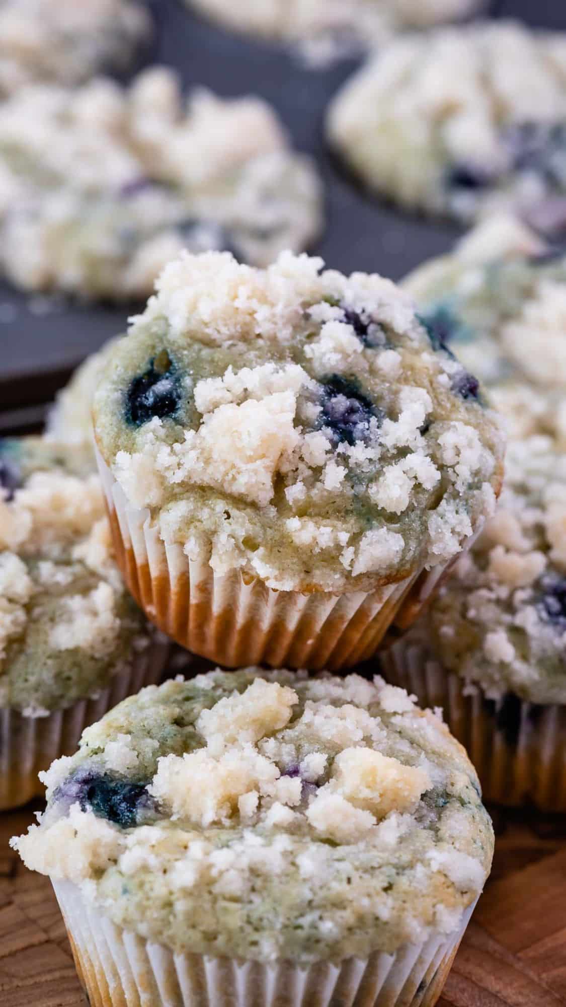 Three blueberry muffins topped with crumbly streusel, arranged in a stack. More muffins are visible in the background on a baking tray.