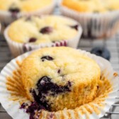 A blueberry muffin with a bite taken out, revealing juicy blueberries inside, sits in an opened paper wrapper. Three more muffins in similar wrappers are in the background on a cooling rack, surrounded by a few scattered blueberries.