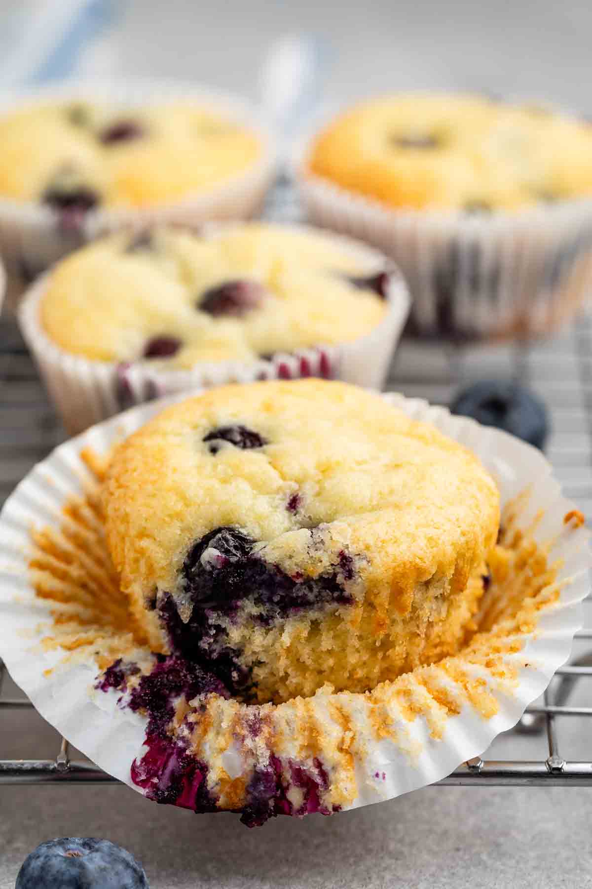 A blueberry muffin with a bite taken out, revealing juicy blueberries inside, sits in an opened paper wrapper. Three more muffins in similar wrappers are in the background on a cooling rack, surrounded by a few scattered blueberries.