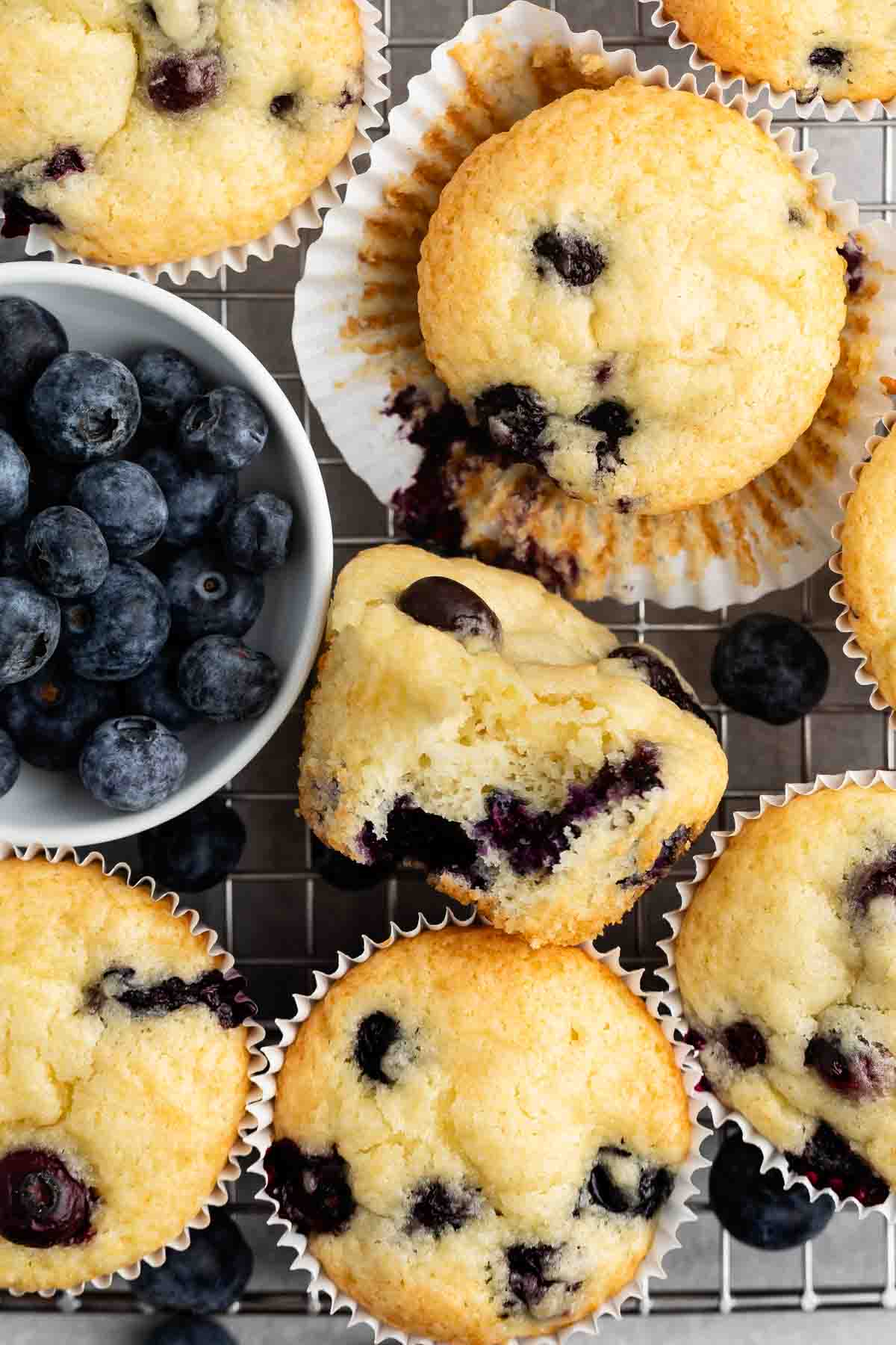 A close-up of freshly baked blueberry muffins on a cooling rack. A bowl filled with fresh blueberries is placed beside the muffins. One muffin is partially eaten, revealing juicy blueberries inside.