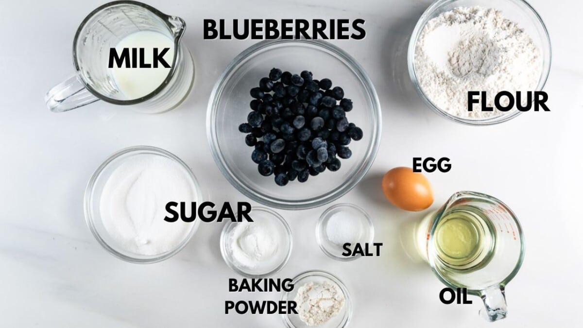 Top-down view of baking ingredients on a white surface: a bowl of blueberries, a pitcher of milk, a bowl of flour, an egg, a bowl of sugar, a small dish of baking powder, a pinch of salt, and a measuring cup with oil.