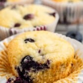 A freshly baked blueberry muffin sits in an opened paper liner. The muffin is golden brown with visible blueberries. In the background, more muffins can be seen slightly blurred, all on a cooling rack.