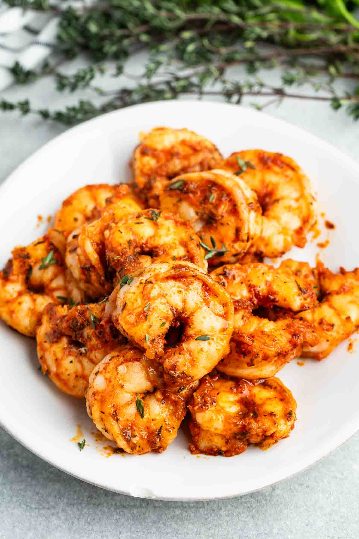 A plate of cooked shrimp seasoned with spices and herbs, garnished with sprigs of thyme. The shrimp have a reddish-orange hue, indicating a flavorful seasoning. The background includes a blurred view of more thyme.