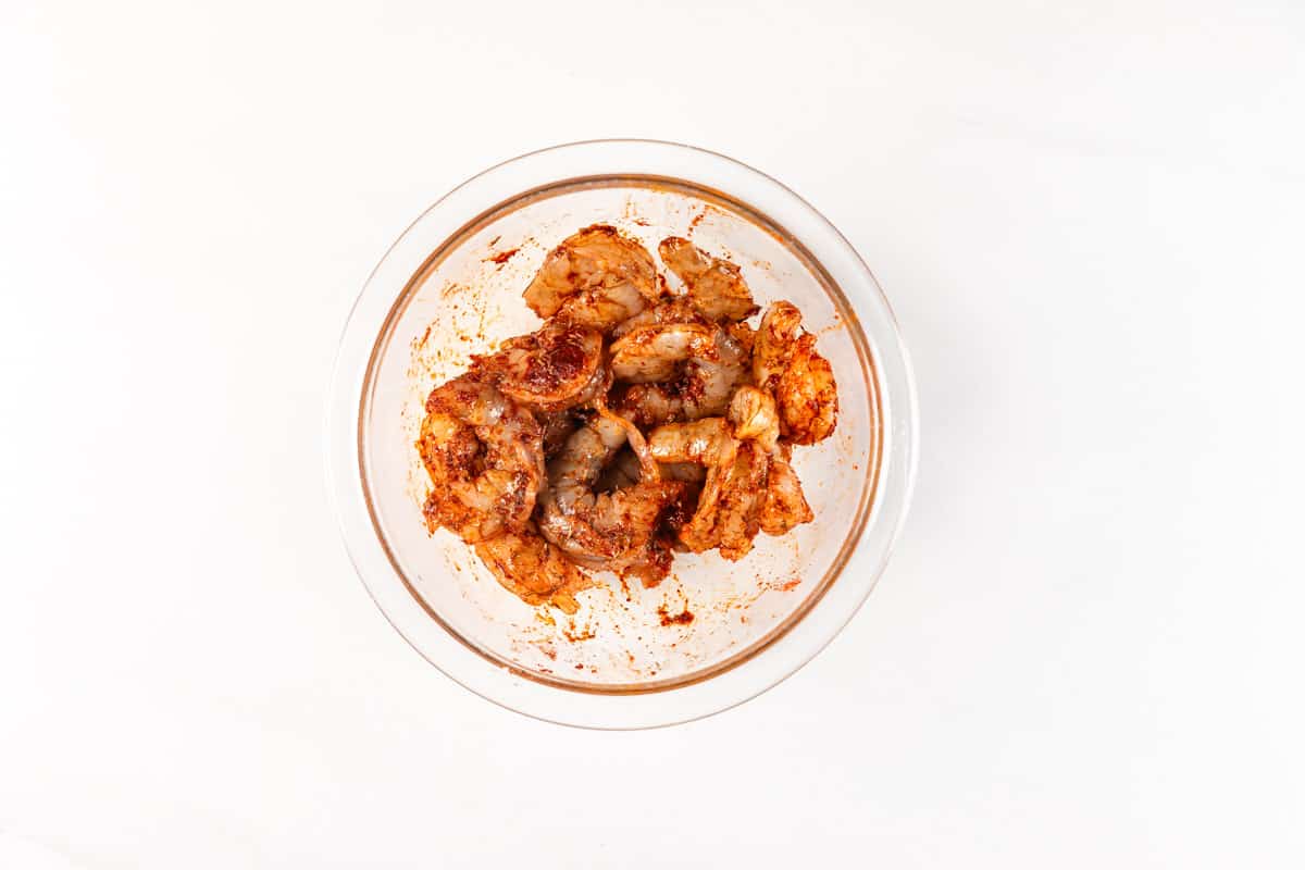 Glass bowl containing raw shrimp marinated in reddish seasoning on a white background.