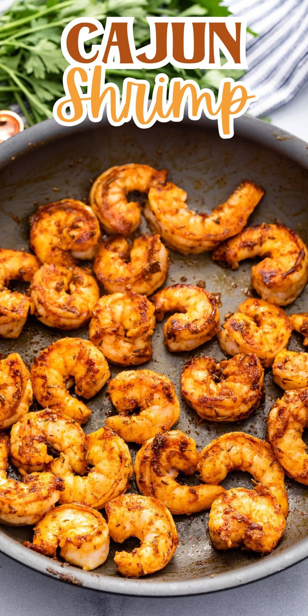 A skillet filled with cooked Cajun shrimp, seasoned with spices, showcasing a rich, reddish-brown color. Leafy greens are partially visible in the background. Text at the top reads Cajun Shrimp.