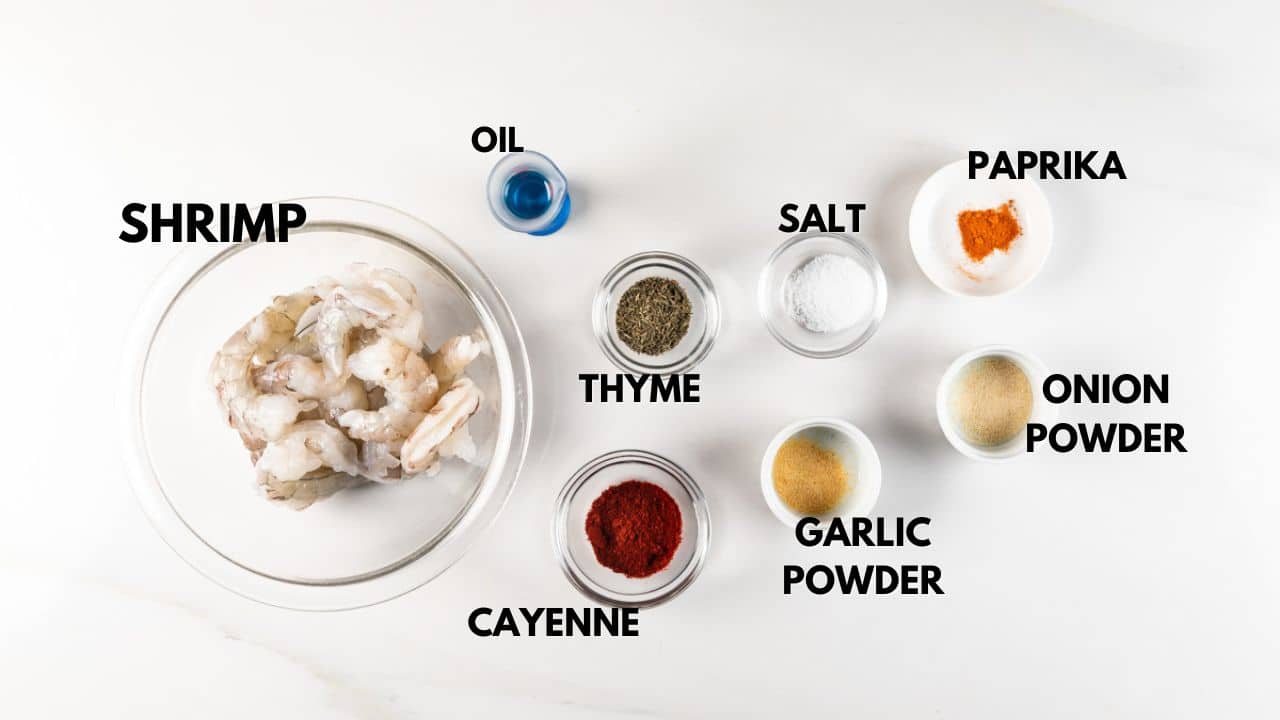 A glass bowl of raw shrimp is surrounded by small containers with spices and liquids, labeled: oil, thyme, paprika, salt, onion powder, garlic powder, and cayenne, arranged on a white surface.