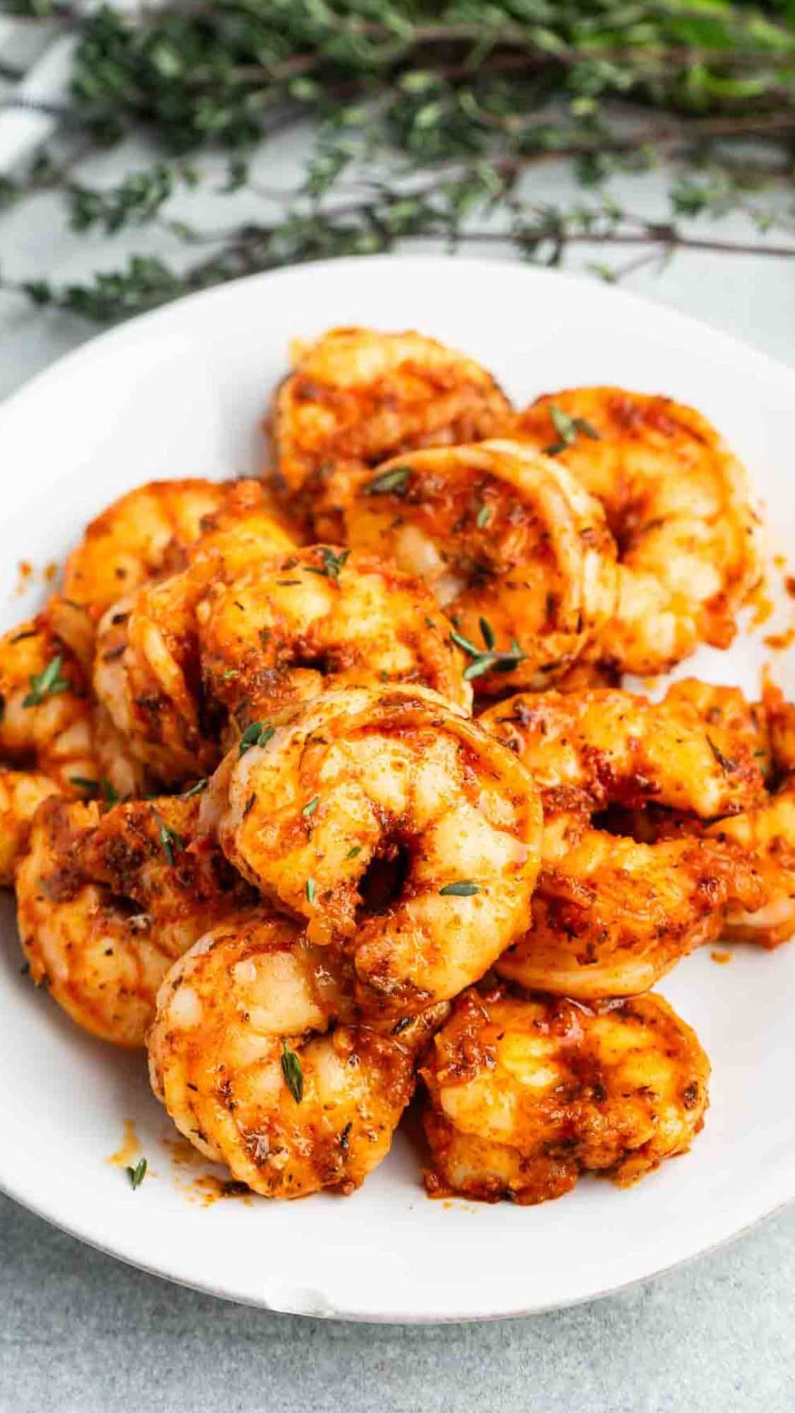 A white plate filled with cooked shrimp seasoned with a reddish spice mix. The shrimp are garnished with small green herbs, and the texture suggests they are freshly cooked. In the background, out of focus, are sprigs of herbs.