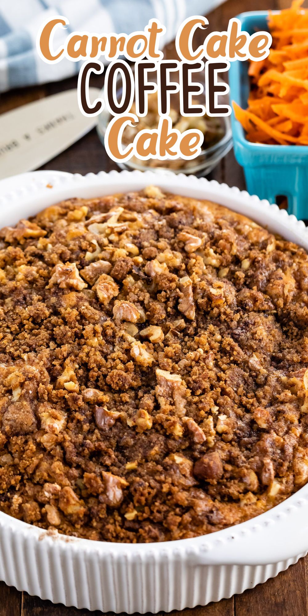 A close-up of a carrot cake coffee cake in a white dish on a wooden table. The cake is topped with a crumbly walnut streusel. In the background, theres a blue basket filled with shredded carrots.