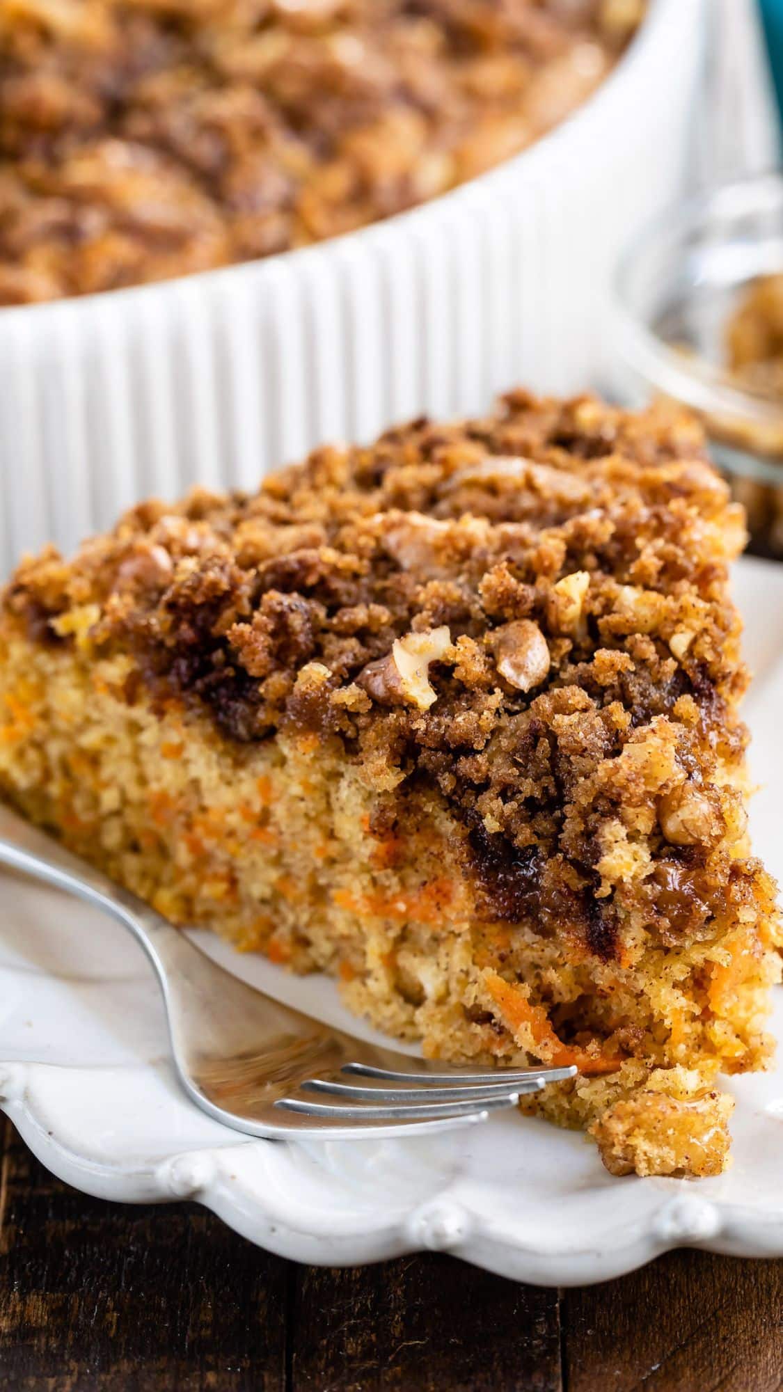 A slice of carrot cake topped with a crumbly streusel and nuts on a white plate. A fork rests beside the cake, with the cakes moist texture and pieces of carrot visible. A white dish with more cake is in the background.