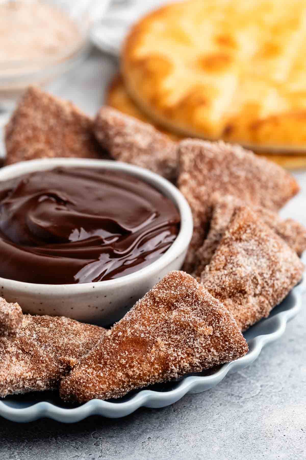 Plate of cinnamon sugar-dusted chips arranged around a bowl of rich chocolate dipping sauce. A golden-brown round pastry is blurred in the background.