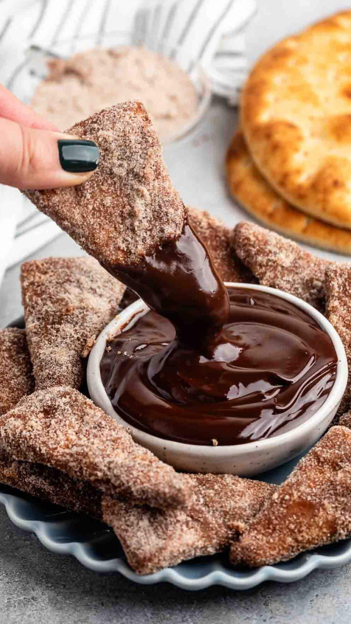 A hand dips a cinnamon-sugar coated tortilla chip into a bowl of rich chocolate sauce. Other chips are arranged around the bowl on a plate, with more chips and tortillas visible in the background.