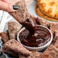 A hand dips a cinnamon sugar-coated tortilla chip into a bowl of rich chocolate sauce. A plate with more cinnamon chips and a stack of round flatbreads are visible in the background on a gray surface.