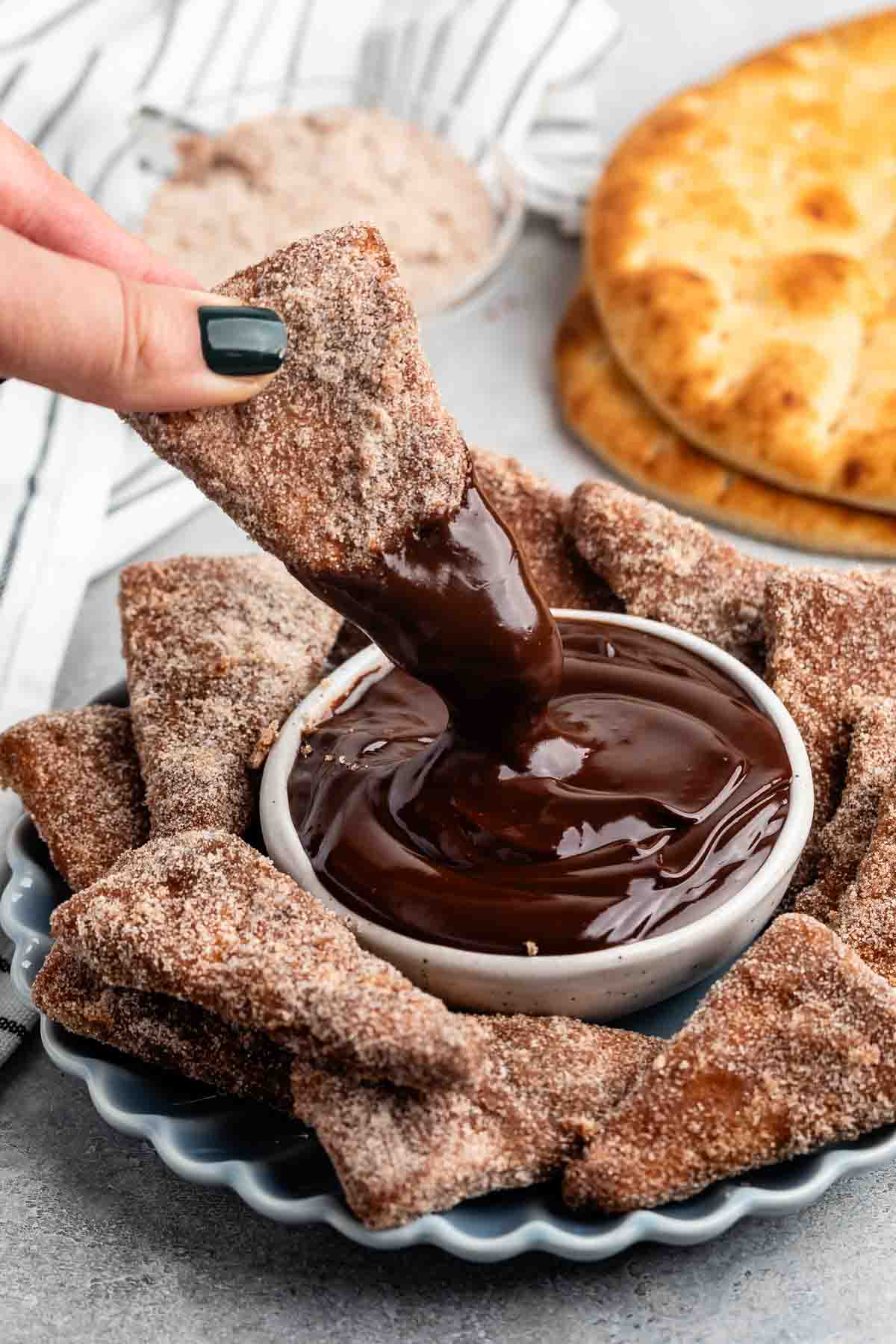 A hand dips a cinnamon sugar-coated tortilla chip into a bowl of rich chocolate sauce. A plate with more cinnamon chips and a stack of round flatbreads are visible in the background on a gray surface.