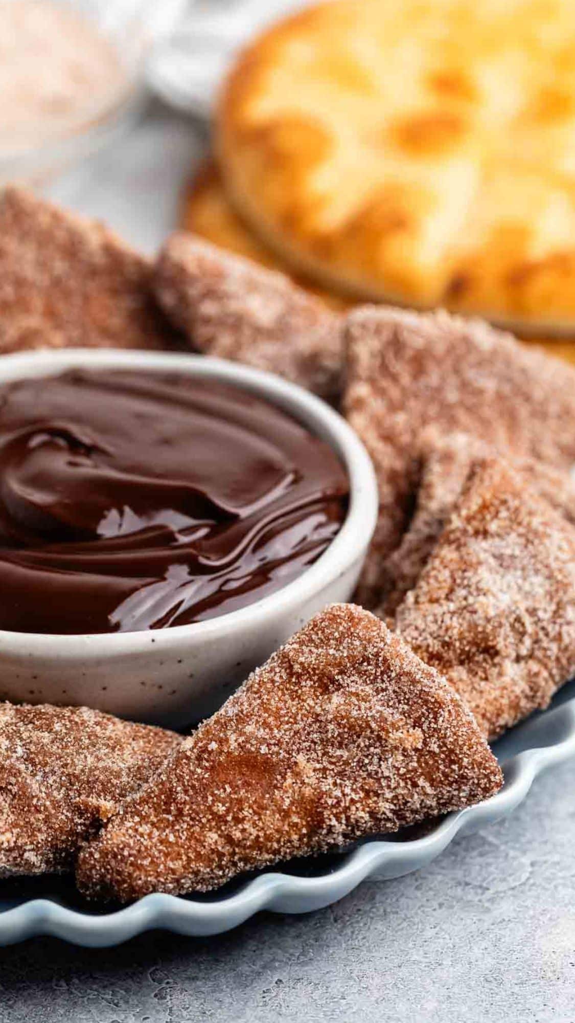 Close-up of cinnamon-sugar coated chips arranged around a small bowl of rich chocolate dipping sauce. A round bread in the background adds a warm, inviting touch to the indulgent snack setting.