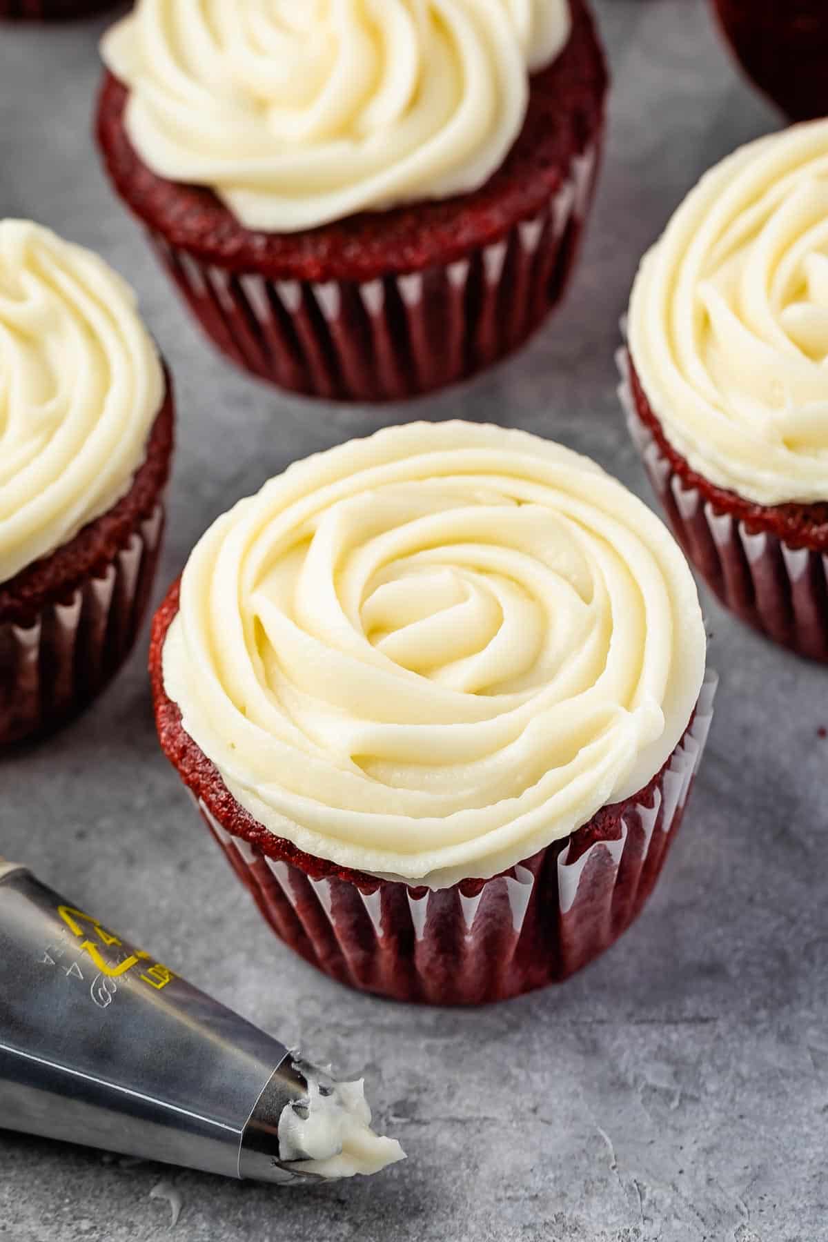 Red velvet cupcakes topped with creamy white frosting, arranged on a gray surface. A piping tip with remaining frosting is nearby, indicating freshly decorated cupcakes.