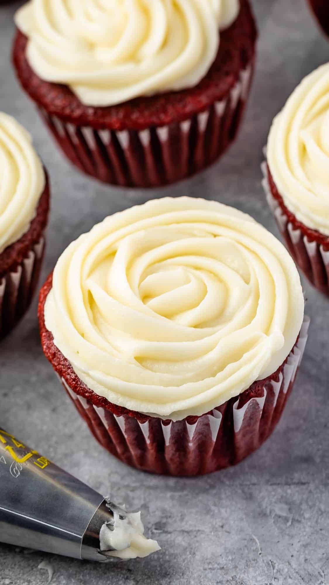 Red velvet cupcakes with cream cheese frosting swirled on top are arranged on a gray surface. A piping bag filled with frosting is nearby, hinting at the decorating process.