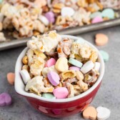 A red bowl filled with a mix of popcorn, pretzels, peanuts, and heart-shaped pastel candies coated in a white glaze. Additional candy hearts are scattered on the gray surface around the bowl. A baking tray with more of the mix is in the background.