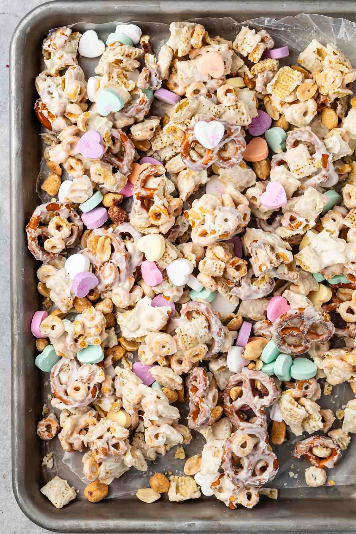 A baking tray filled with a colorful snack mix, including pretzels, cereal pieces, peanuts, and pastel-colored candy hearts. The items are coated in a light sugary glaze, creating a sweet and crunchy treat.