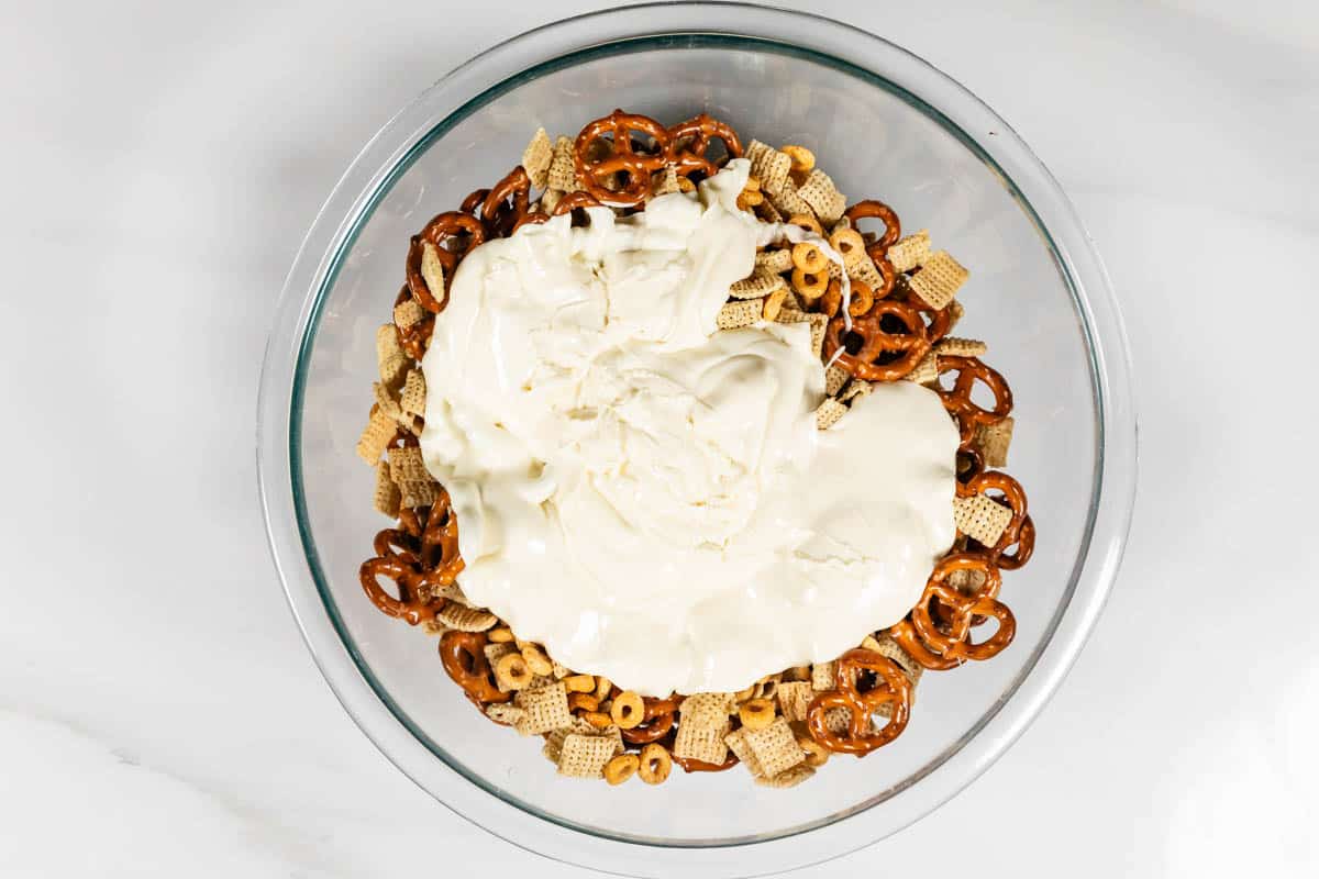 A glass bowl contains a mixture of pretzels, cereal squares, and cereal loops topped with a thick layer of white melted chocolate. The bowl is placed on a white surface.