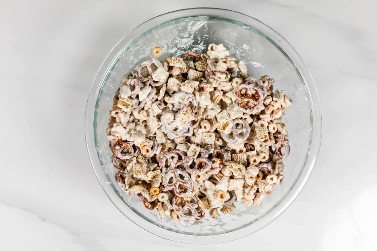 A glass bowl filled with a snack mix that includes cereal pieces, pretzels, and possibly nuts, all coated in a white, sugary glaze, placed on a light marble surface.