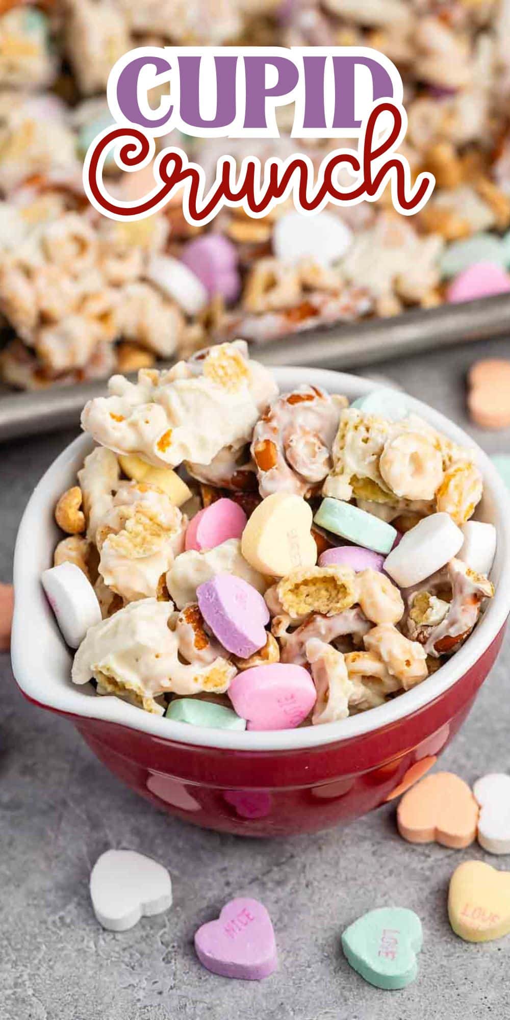 A close-up of a red bowl filled with Cupid Crunch, a snack mix of popcorn, pretzels, cereal, and colorful candy hearts, topped with a white coating. Loose candy hearts are scattered around the bowl on a gray surface.