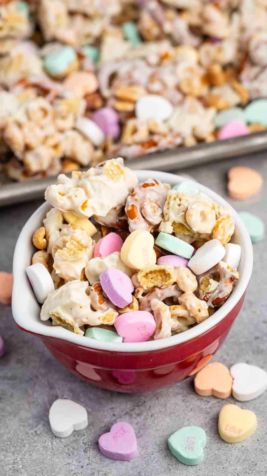 A red bowl filled with a mix of popcorn, nuts, and colorful heart-shaped candies coated in a white drizzle. More candies and similar snack mix are scattered on a baking tray in the background.