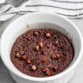 A microwave chocolate mug cake topped with chocolate chips in a white ramekin. A striped dish towel and a spoon are in the background on a gray surface.