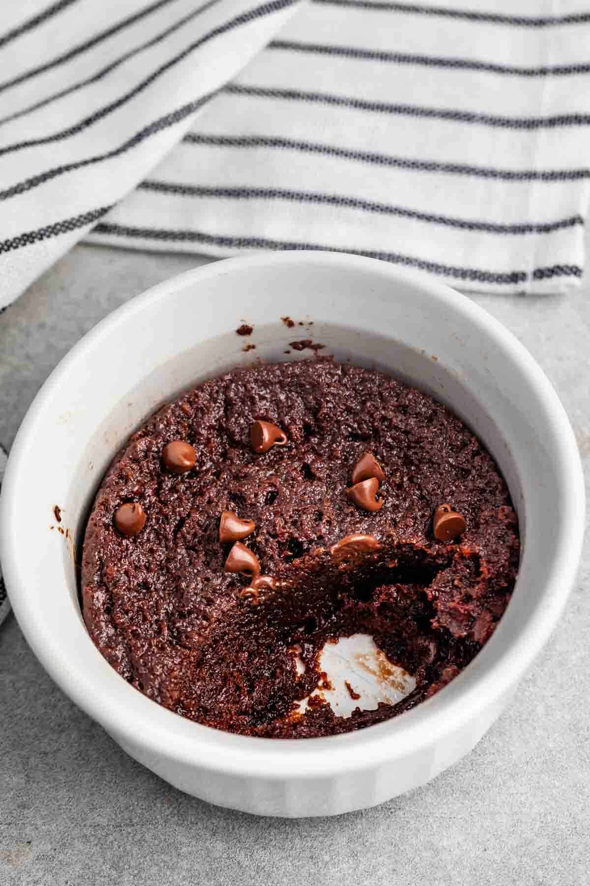 A small white ramekin filled with a chocolate mug cake topped with a few chocolate chips. A portion of the cake has been eaten, revealing its moist texture. The ramekin is on a gray surface with a striped cloth in the background.