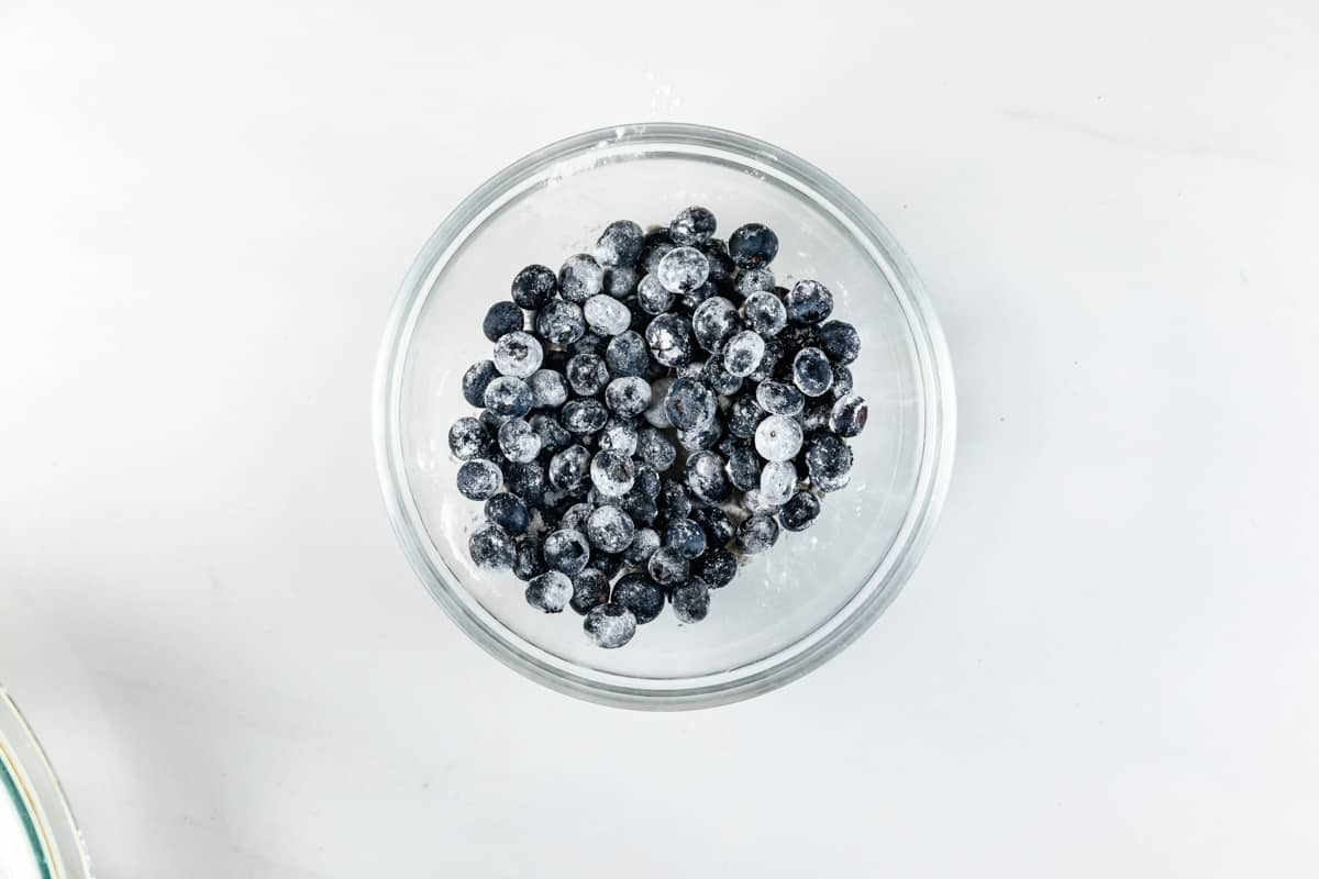 A clear glass bowl filled with blueberries, lightly dusted with flour, placed on a white surface.