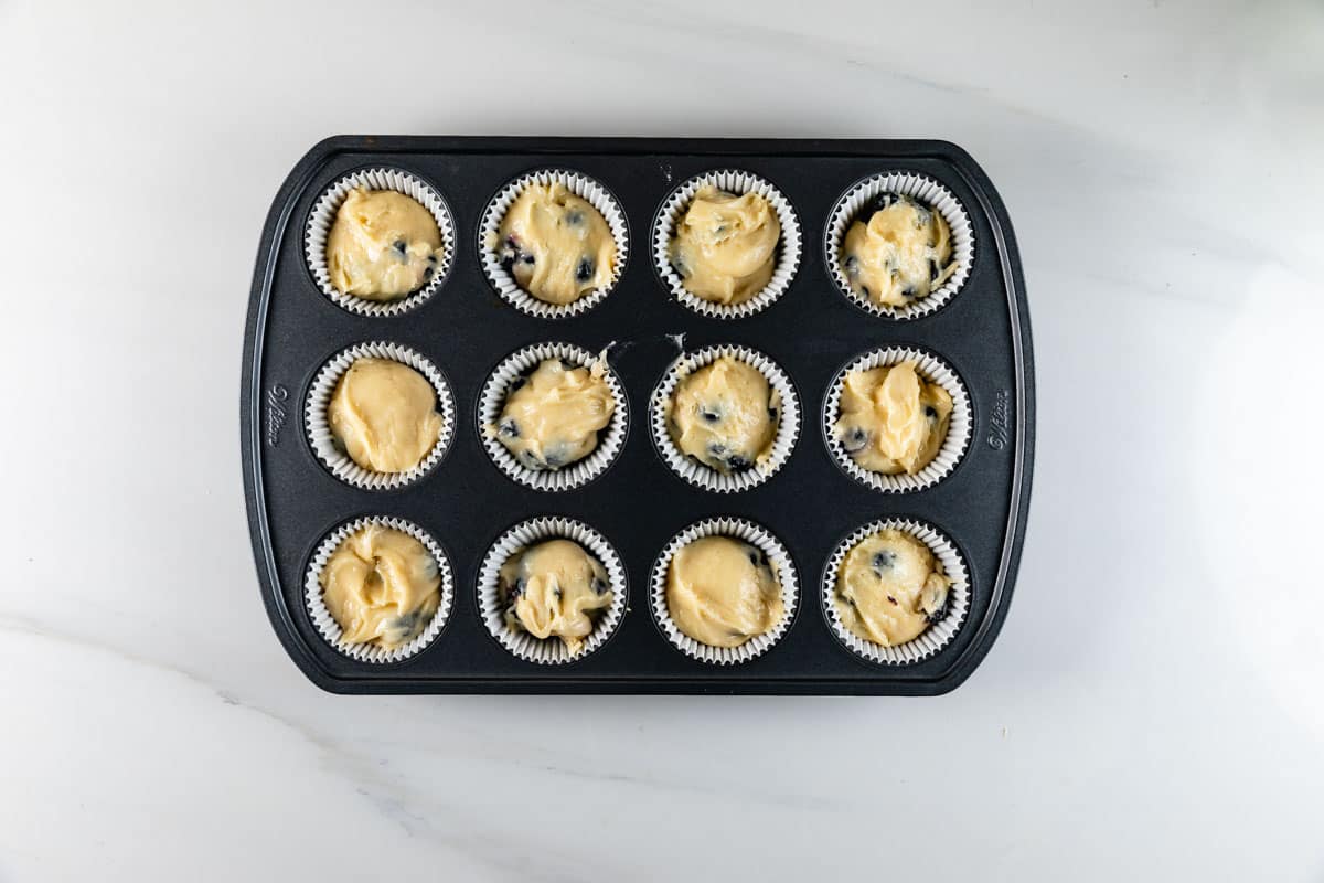 A muffin tin with twelve paper liners, each filled with raw muffin batter containing visible blueberries, placed on a light countertop.